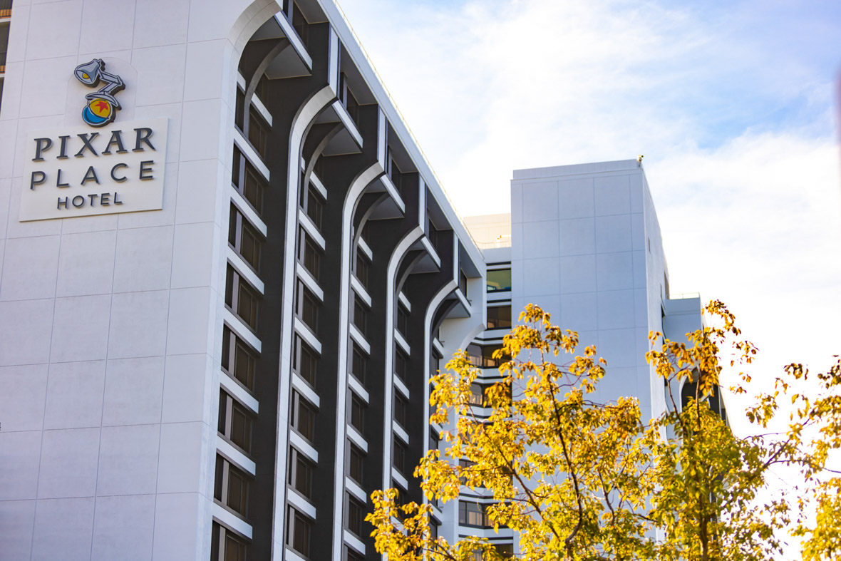 An exterior image of Pixar Place Hotel shows the 15-story tower with the hotel’s name on one side. The branches and yellow leaves of a tree are in the lower right foreground.