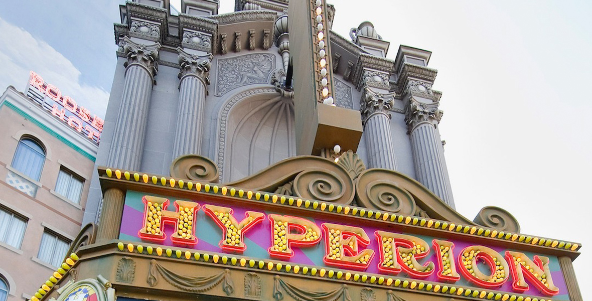 An exterior shot of the Hyperion Theater at Disney California Adventure Park. It is daytime and the top of the marquee is visible, showcasing large, capitalized, serif letters spelling out "Hyperion" in yellow light bulbs surrounded by a red trim. Towering above the marquee are grey, stone columns dotted with blinking white lights. A painted city background is barely visible to the left of the theater entrance.
