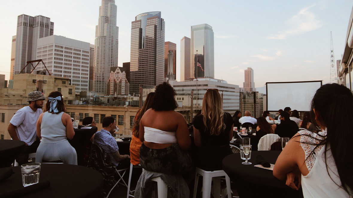 A crowd sitting in lawn chairs in front of a large movie screen with a city skyline in the background.