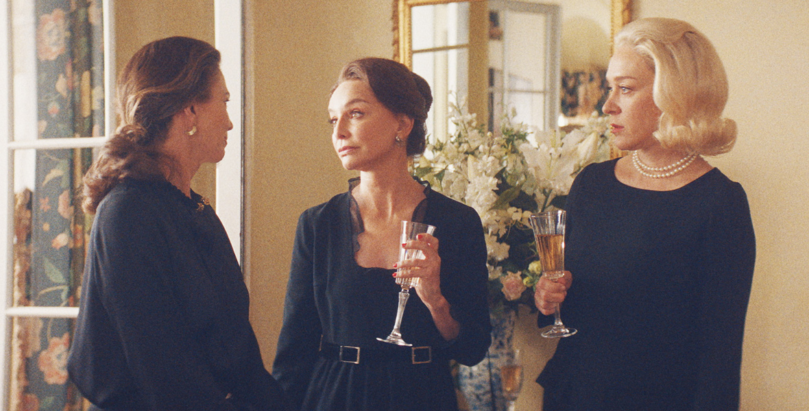 In a scene from the television series Feud: Capote vs. The Swans, from left to right, Diane Lane, Calista Flockhart, and Chloë Sevigny stand indoors conversing. They are in black dresses, with Flockhart and Sevigny seen holding drinks as they gaze toward Diane.